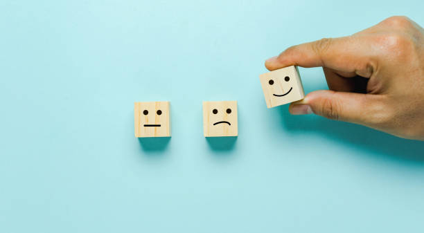 Top view of  Hand holding of Happy icon on cube blocks about rating feedback of survey from customer. Business annual survey concept. Many sad or happily on wooden on blue paper. stock photo