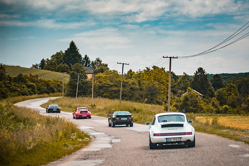 Porsches driving in the group on the open mountain road. Different models are on the photo, from the 911,944, 993 and Boxter models. This photo represent enthusiast driving during weekend and enjoying in mountain road and ride. Photo have vintage look and was taken on the cloudy day.