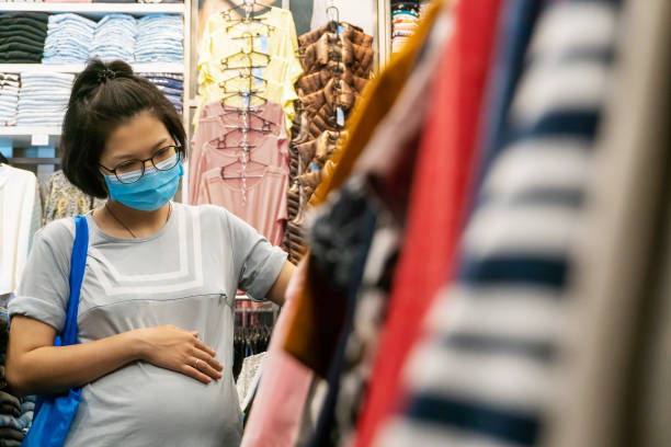 donna incinta asiatica con maschera facciale per proteggere il virus corona mentre sceglie nuovi abiti nel negozio di abbigliamento in un centro commerciale. - human pregnancy clothing shopping retail foto e immagini stock