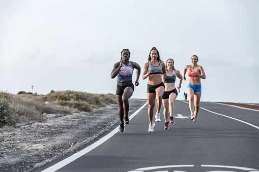 Focused diverse friends in sportswear running on asphalt road towards camera during outdoor training against gray sky