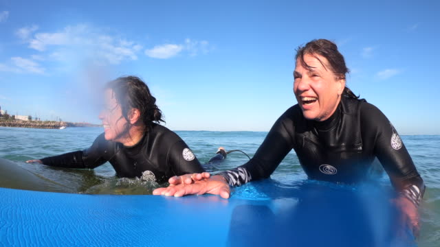 Handheld close-up of two mature caucasian women lying down on their surfboards and talking in the sea.