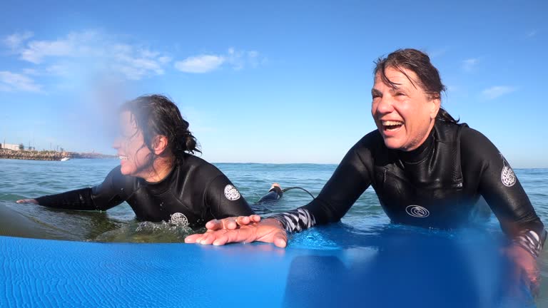 Handheld close-up of two mature caucasian women lying down on their surfboards and talking in the sea.