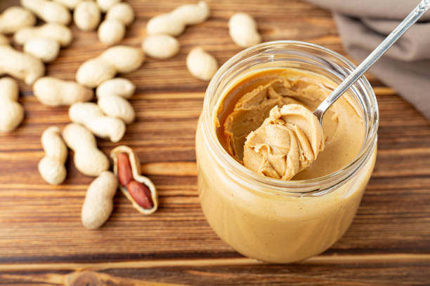 istock Creamy peanut paste in open glass jar, peanut butter in spoon. Peanuts in the peel scattered on the brown wooden table with copy space for cooking breakfast. Vegan food concept. 1248562639