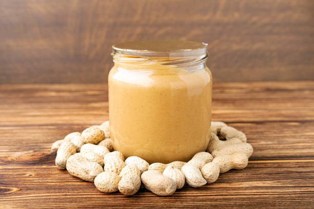 istock Creamy peanut paste in open glass jar, peanut butter spoon, peanuts in the peel scattered on the brown wooden table with copy space for cooking breakfast. Vegan food concept. 1248562641