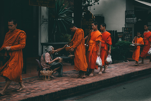 Thai monks  are getting food donations  at  ceremony of Makha Bucha Day in Sangkhla Buri in northwest of Kanchanaburi province of Thailand in early morning close to sunset. People are wearing mostly traditional clothing and are standing in main street leading down to Mon Bridge and lake. People are waiting to give food donations to monks when they pass along street down to bridge. View from behind men