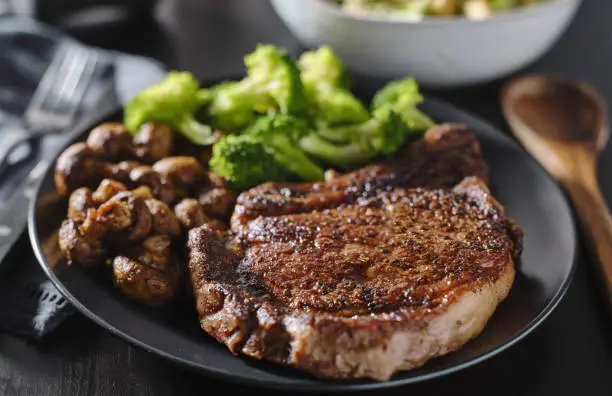 seared ribeye steak with broccoli and sauteed mushrooms on black plate.