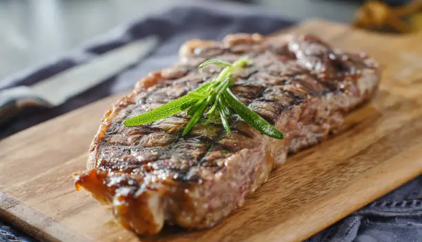 grilled new york strip steak resting on wooden cutting board with rosemary garnish