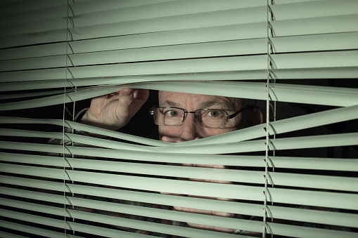Mature man with glasses peeking through window by unfolding blinds