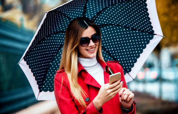 istock Young woman holding umbrella and typing a message on the smartphone while waiting for a boyfriend. Lifestyle, technology concept 1248594516