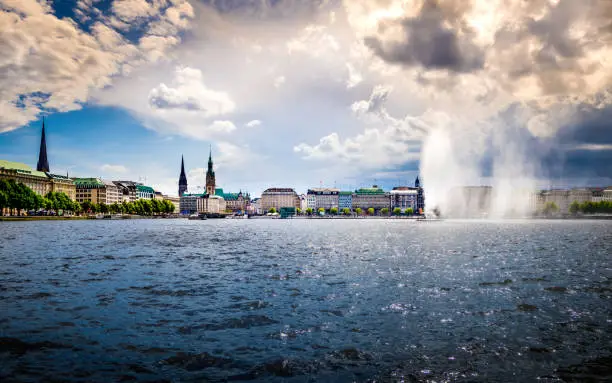 idyllic horizontal panorama of the binnenalster waterfront in hamburg old town with view to the jungfernstieg with town hall and majestic church towers in the background under dramatic cloudscape