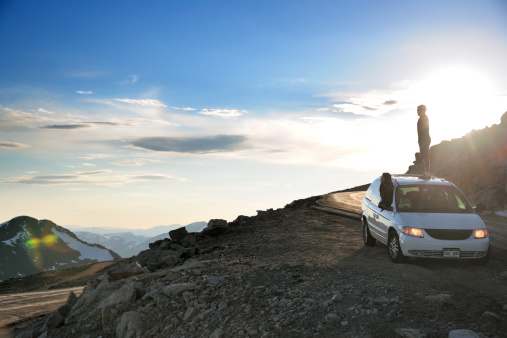 Man (35 years old) standing on top of his minivan with his family and wife (35 years old) leaning out the window as they admire the scenic view from the top of a mountain.