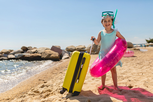 Little girl standing on a sandy beach while holding an inflatable ring near her suitcase wearing a snorkel