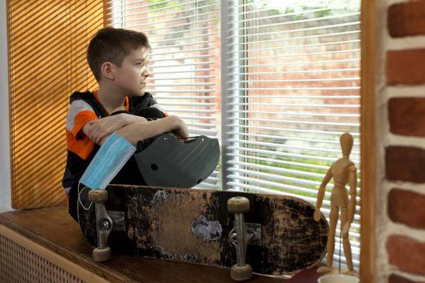 Sad boy is sitting near the window with his skateboard and medical safety mask during the quarantine stock photo