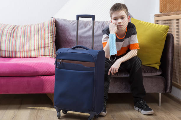 Sad boy is sitting at home near his suitcase with safety mask in his hand. stock photo