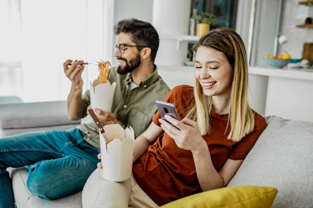 Home delivery of food for young couple A young couple is eating pasta at home and smiling, a young woman is holding a mobile phone food delivery stock pictures, royalty-free photos & images