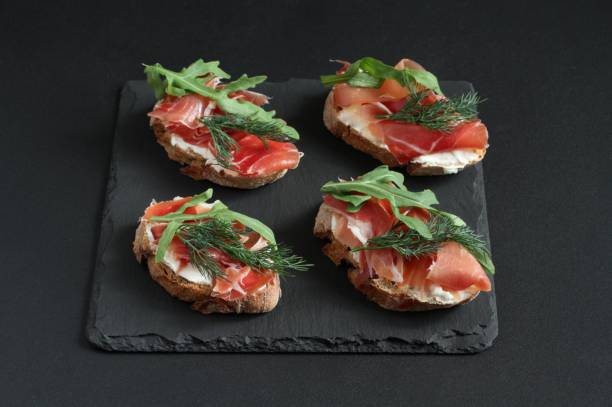 istock Buckwheat toasts with goat cheese, prosciutto and arugula lying on a black stone board on a black table. Closeup 1248610770