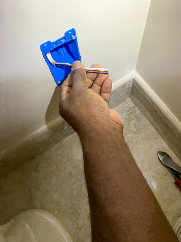 Unrecognizable African American electrician is installing an electrical outlet in a bathroom wall.