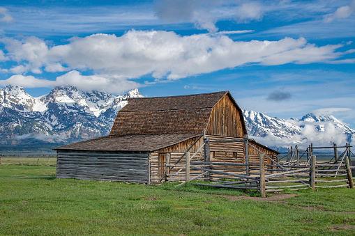 A deserted historic cabin in the meadow of the Grand Teton National Park near the town of Jackson Wyoming, USA with the snow-capped mountain range and blue sky with clouds in the background during summer.