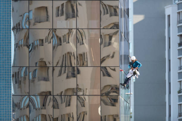 Image of a young skyscraper window washer wearing safety equipments and cleaning a glass curtain wall building facade
