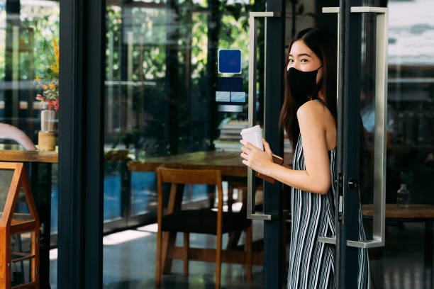 giovane bella donna asiatica che indossa una maschera chirurgica mentre lascia una caffetteria. attraente cliente femminile che acquista caffè e tiene una tazza di caffè usa e getta. prevenzione covid-19 - starbucks women walking restaurant foto e immagini stock