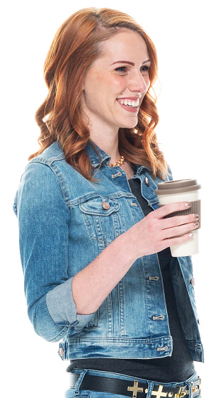Side view of aged 20-29 years old who is beautiful with long hair caucasian young women standing in front of white background wearing denim jacket who is laughing and holding coffee cup