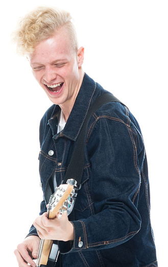 Profile view of aged 20-29 years old with blond hair caucasian young male guitarist standing in front of white background wearing leather jacket who is happy and holding electric guitar