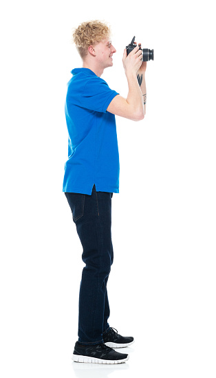 Full length of aged 20-29 years old with blond hair caucasian male tourist standing in front of white background wearing polo shirt who is laughing who is photographing and holding camera