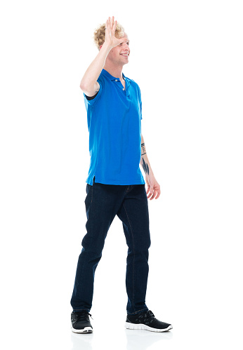 Full length of aged 20-29 years old with curly hair caucasian young male standing in front of white background wearing jeans who is laughing and greeting and showing high-five