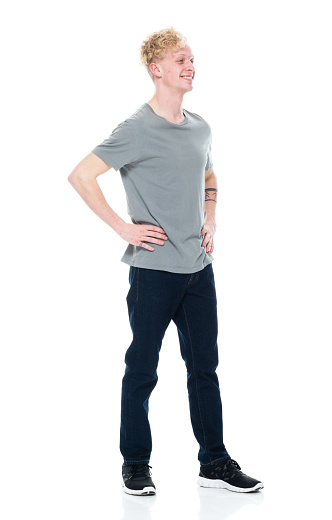 Side view of aged 20-29 years old with curly hair caucasian young male standing in front of white background wearing jeans who is showing cool attitude with hand on hip