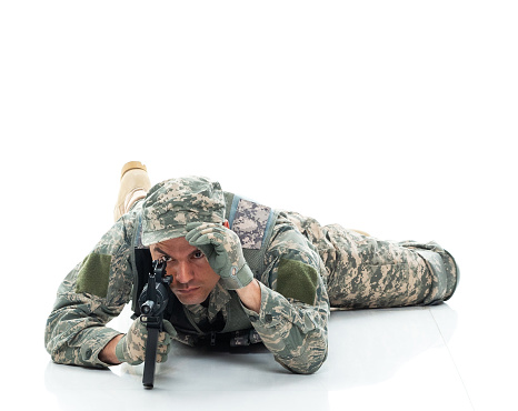 Front view of aged 30-39 years old who is tall person with black hair caucasian male army soldier lying down in front of white background in the military wearing military uniform who is serious and showing patriotism who is shooting a weapon and holding weapon and using gun