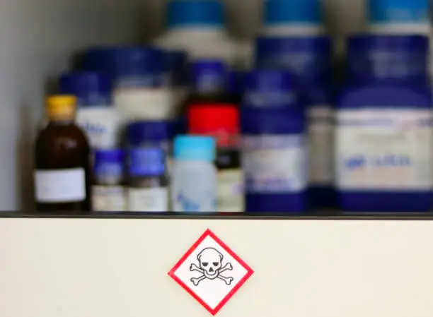 Photo of Chemical Hazard Sign pictogram, Globally Harmonized System of Classification and Labelling of Chemicals (GHS) Toxic category with container bottle on shelf cabinet, selective focus blurred background.