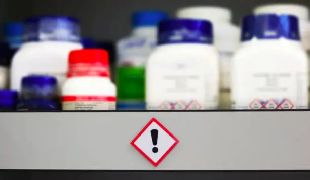 Photo of Chemical Hazard Sign pictogram, Globally Harmonized System of Classification and Labelling of Chemicals (GHS) Harmful Irritation category with container bottles on shelf cabinet, blurred background.