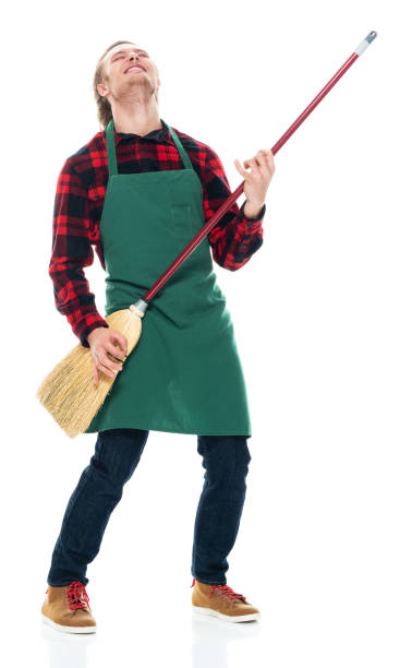 Caucasian young male cleaner bending in front of white background wearing apron and holding broom One person of with blond hair caucasian young male cleaner bending in front of white background wearing apron who is singing who is cleaning and holding broom air guitar stock pictures, royalty-free photos & images