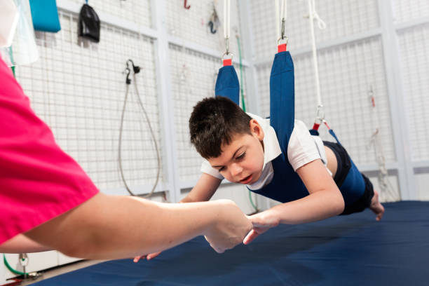 disabled caucasian child doing physiotherapy rehabilitation on elastic cords with physiotherapist, osteopath during coronavirus. mask, cap and glove protections