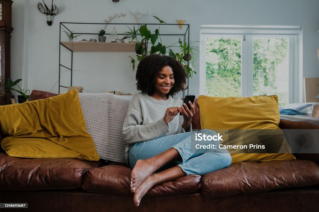 Beautiful woman with afro on smartphone at home in the lounge Beautiful mixed face female on her cell phone or smartphone browsing and smiling while sitting on the couch in her lounge at home Domestic Life Stock Photo