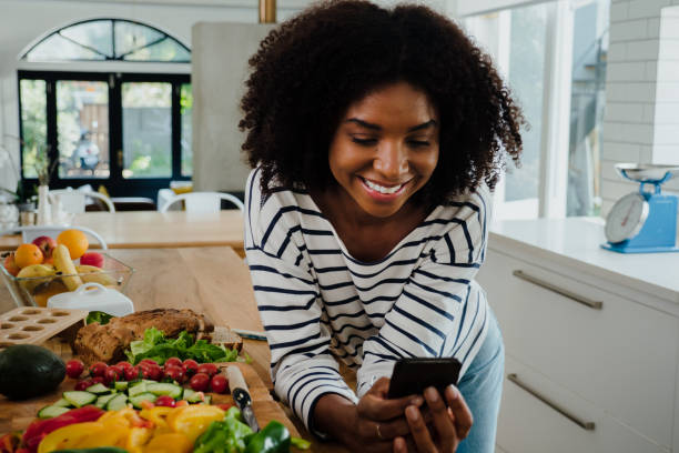 Beautiful woman with afro in the kitchen browsing mobile device Beautiful woman with afro in the kitchen browsing smartphone and smiling, mixed race scrolling photos stock pictures, royalty-free photos & images