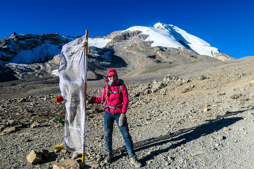 Woman standing next to a prayer flag on her way to the top of Thorung La Pass, Annapurna Circuit Trek Nepal. Harsh and barren landscape around. Clear and blue sky. Snow capped mountains. Early morning