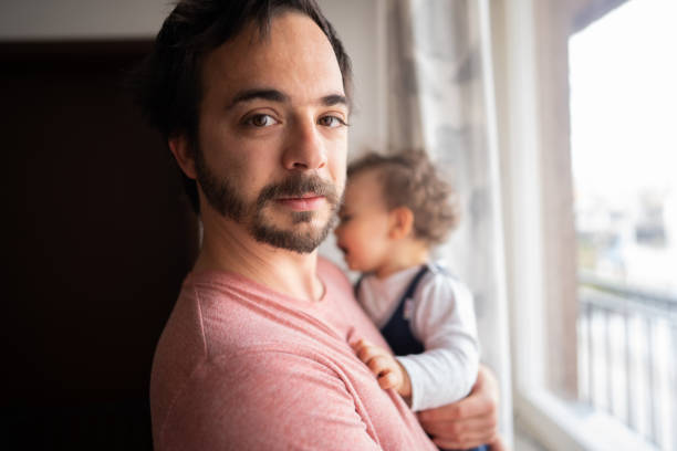 Man standing near window and holding his son in arms, looking at camera