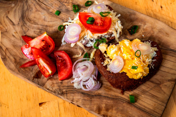 Top view of two toasts with fried eggs with onion, grated cheese and tomatoes on kitchen board of olive wood on table.