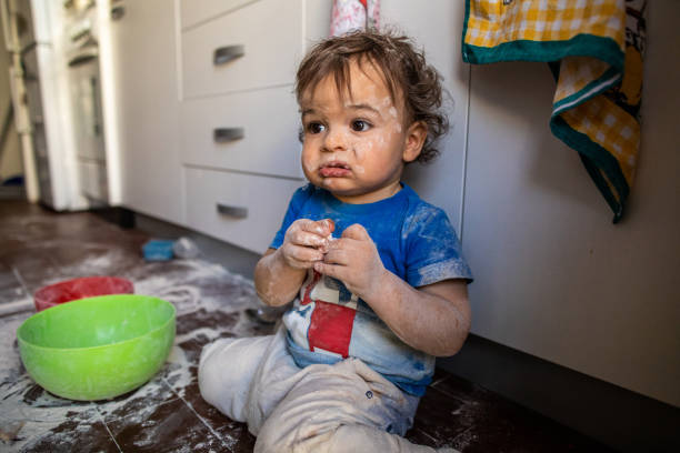 Cute little boy sitting on floor with mess he made with flour around him at kitchen