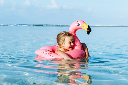 4 years old little girl, wearing necklace made of seashells, swimming in a tropical ocean on inflatable pink flamingo. Blue sky with few fluffy clouds on the background.