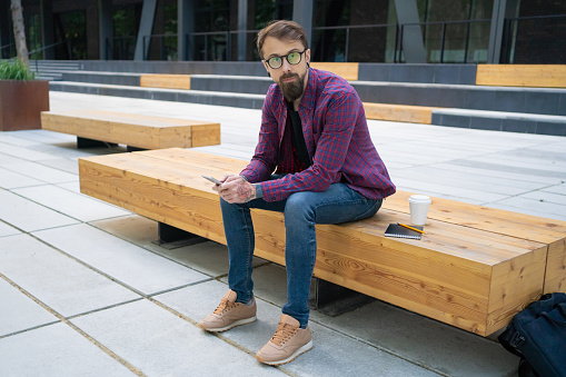 Handsome man sitting on wooden bench with phone. Pencil, notebook and takeaway coffee near him. Stairs and building on background. Working outside and freelance concept