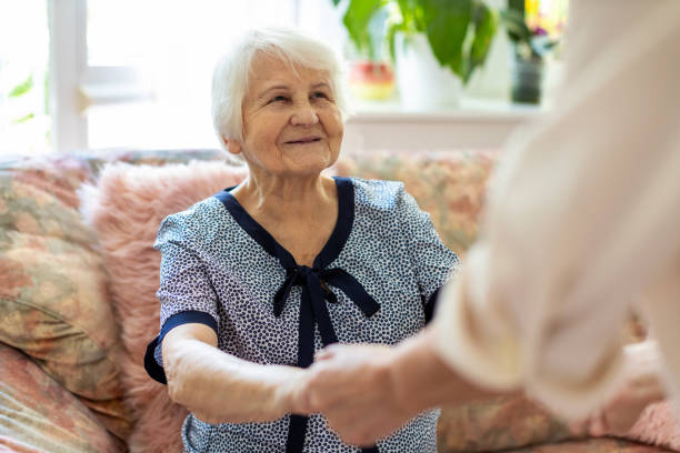 female home carer supporting old woman to stand up from the sofa at care home - alzheimer imagens e fotografias de stock