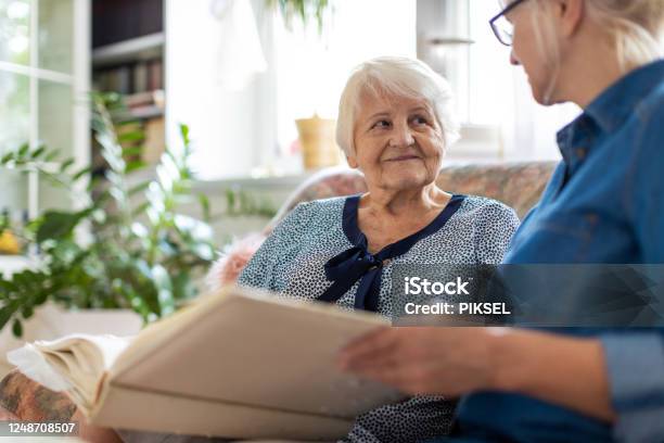 Senior Woman And Her Adult Daughter Looking At Photo Album Together On Couch In Living Room Stock Photo - Download Image Now