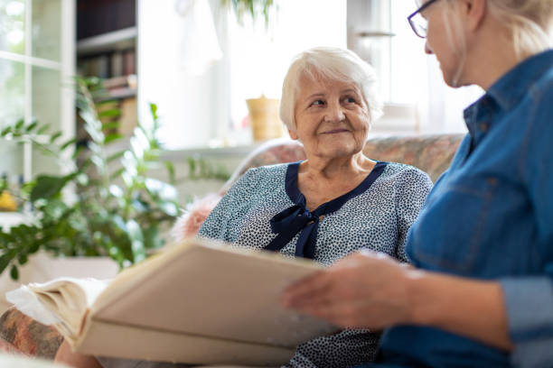 старшая женщина и ее взрослая дочь смотрят на фотоальбом вместе на диване в гостиной - dementia стоковые фото и изображения