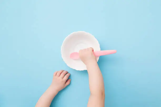 Baby hand putting pink plastic spoon in empty bowl and waiting food on light blue table background. Pastel color. Closeup. Point of view shot. Top down view.