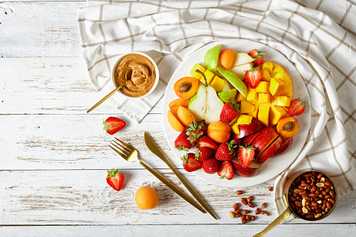 Colorful fruit dessert: platter of strawberries, mango, apples, pears, apricots, with peanut butter and peanuts on a white plate on a white wooden table, top view, flat lay, copy space
