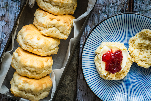 A freshly baked scone on a blue plate smothered in clotted cream and raspberry jam. There are five scones in a metal tin lined with greaseproof paper beside the plate. The background is a painted wooden surface. The paint is old and flaking from the surface. The focus is on the jam.