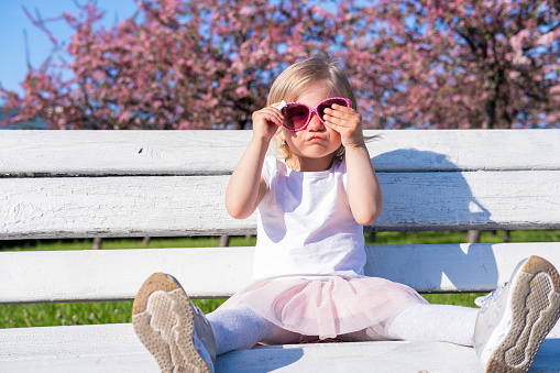 Funny little baby girl sitting on white bench in city park in pink glasses at summer, looking at camera. Happy blonde child playing having fun outdoor