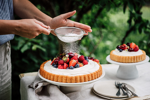 Close-up of a woman hands sifting powdered sugar on a cheesecake. Female preparing fresh fruits and berries cake outdoors in a garden.